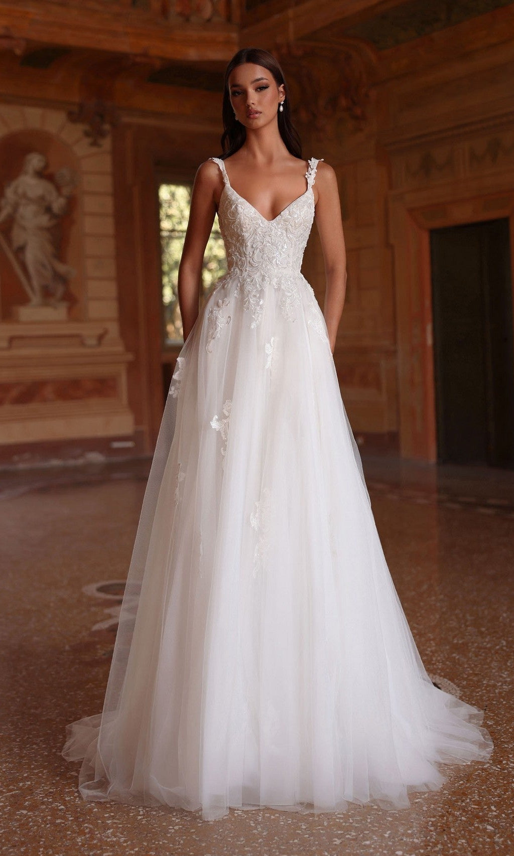 A woman wearing the Alyce 7124 long ivory dress—sleeveless, floor-length A-line with lace appliques—stands indoors in an ornate room decorated with classical art and warm lighting.